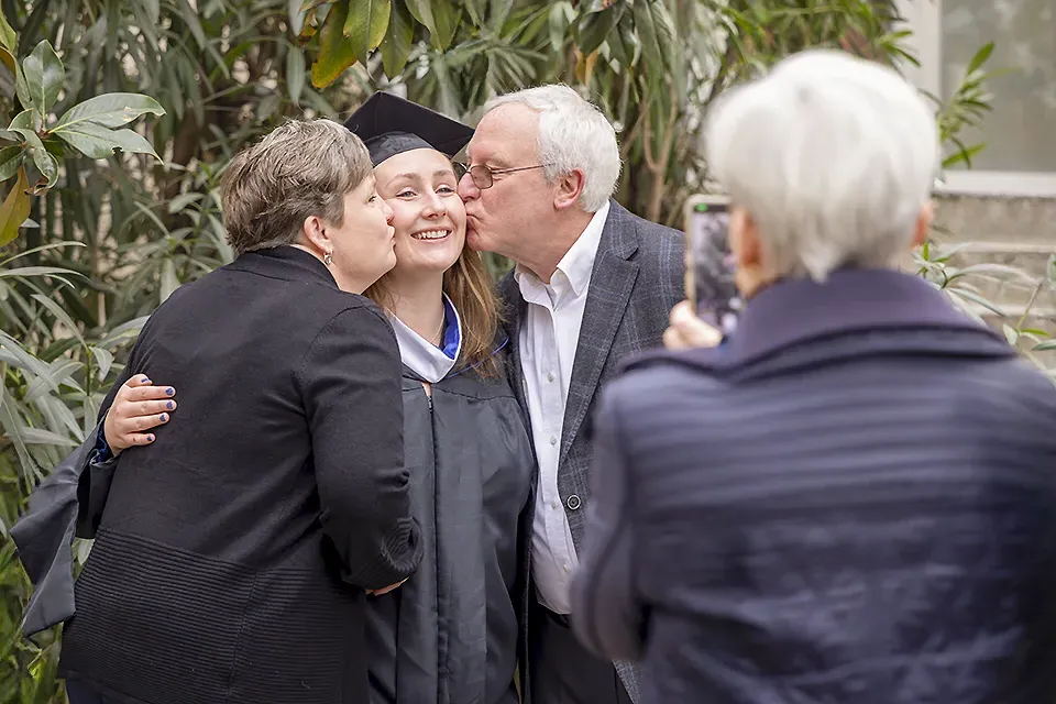 A woman hugs a participant in a graduation ceremony and looks up at the sky, while others mill around the area.